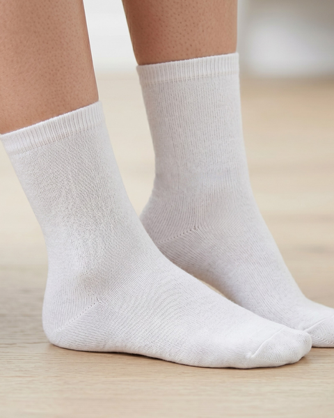 A close-up view of a person's lower legs and feet wearing plain white, fine-knit cotton crew socks. The person is standing on a light-colored wooden floor. The background is softly blurred.