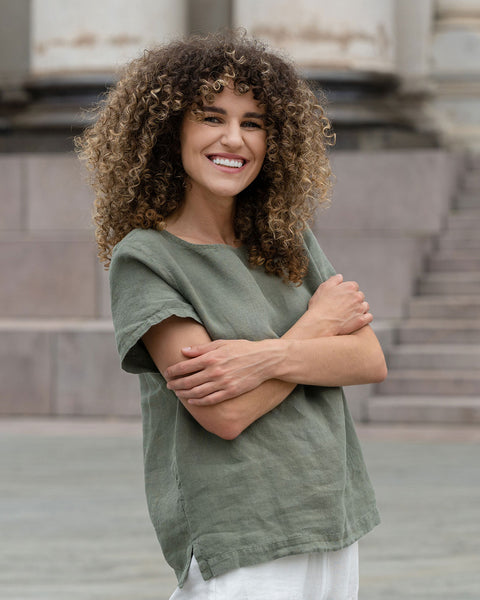Beautiful woman with a curly hair in the city, posing wearing stone green linen blouse Emma and pure white linen pants Dakota.