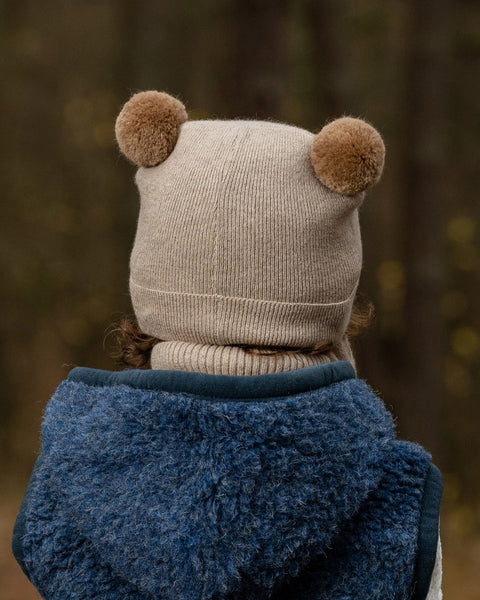 The back of a young child&#39;s head and shoulders are shown, with the child wearing a beige knit hat with two brown pom-pom ears. They also have a beige turtleneck and a dark blue fuzzy vest on, with a blurred outdoor background.