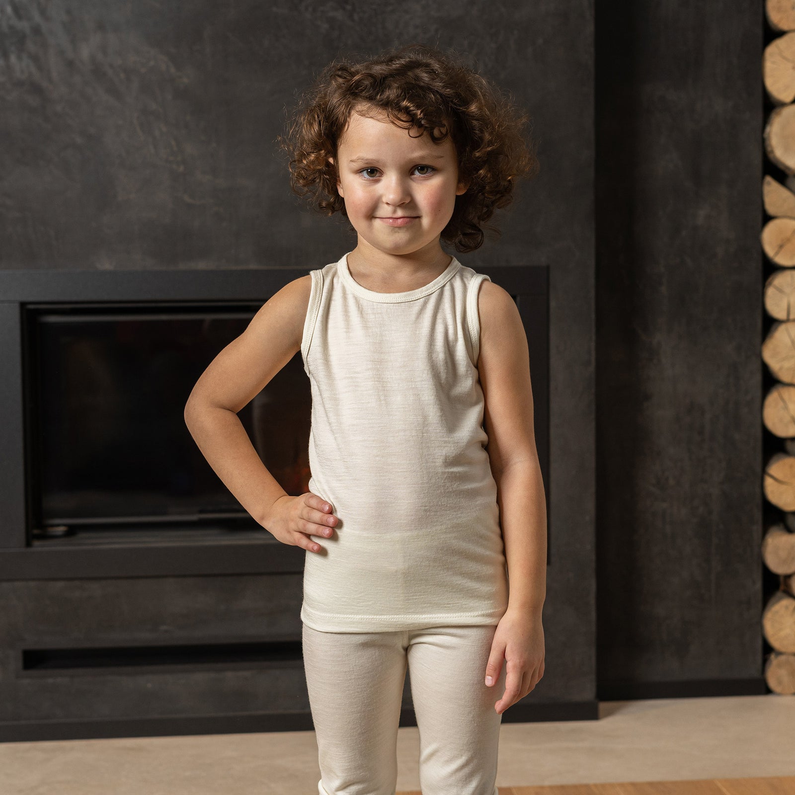 A young child with light curly hair in pigtails is sitting on a white surface against a plain white background. The child is wearing a menique sleeveless top and matching leggings. They are looking directly at the camera with a pouty expression, and their bare feet are visible.