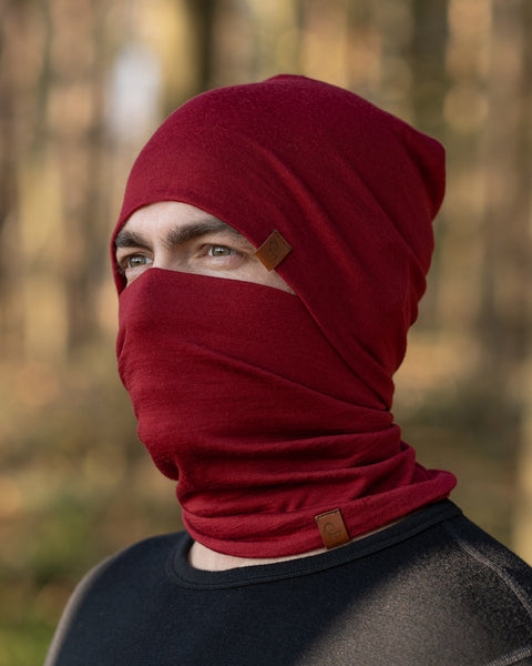 A man, standing outside in a forest during fall season, wearing the Menique Men&#39;s Merino Beanie and neck Gaiter Set in the color Royal Cherry.