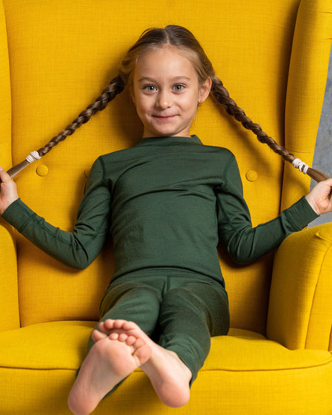 A girl smiling at the camera, wearing the menique Kids&#39; Long Sleeve Crew 160 Merino in Dark Green color. She is sitting in a yellow ottoman, holding her braids with her hands to the sides.