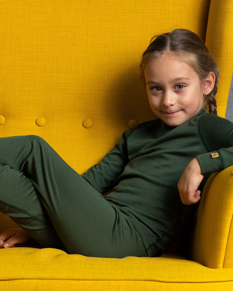 A girl sitting on a yellow armchair, wearing the menique Kids&#39; Merino 160 Pants in Dark Green color.